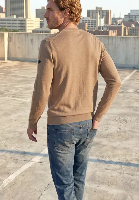 Trui met ronde hals van puur katoen antique beige Een man in een bruine trui en spijkerbroek staat op een parkeerdak, uitkijkend over de stad.