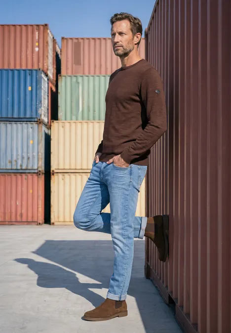 Trui met ronde hals van puur katoen maroon brown Een nonchalante man leunt ontspannen tegen een felgekleurde zeecontainer, gekleed in een bruine trui en spijkerbroek.