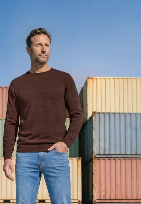 Trui met ronde hals van puur katoen maroon brown Een man in een bruine trui en spijkerbroek staat voor gekleurde containers.