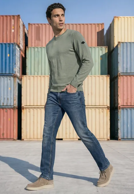Trui met ronde hals van puur katoen willow green Een man in een groen shirt met lange mouwen, jeans en lichte schoenen staat voor kleurrijke zeecontainers.