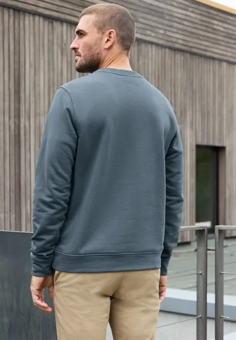 Un homme vu de dos en sweat gris-bleu et pantalon beige devant un bâtiment en bois.