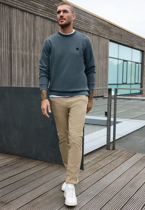 Un homme en sweat gris et pantalon beige marche sur une terrasse en bois devant un bâtiment moderne.