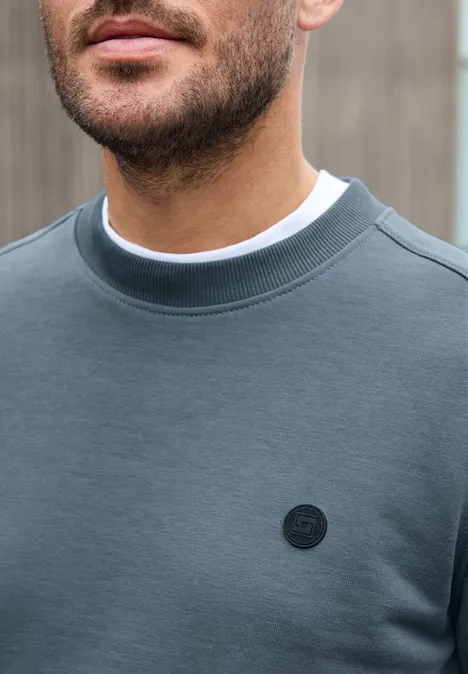 Un homme porte un sweat-shirt gris sobre avec un petit logo rond noir sur la poitrine, devant un arrière-plan flou.