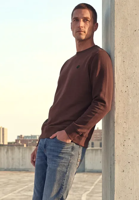 Un homme décontracté appuyé contre un pilier sur un parking en hauteur, la ville en arrière-plan.