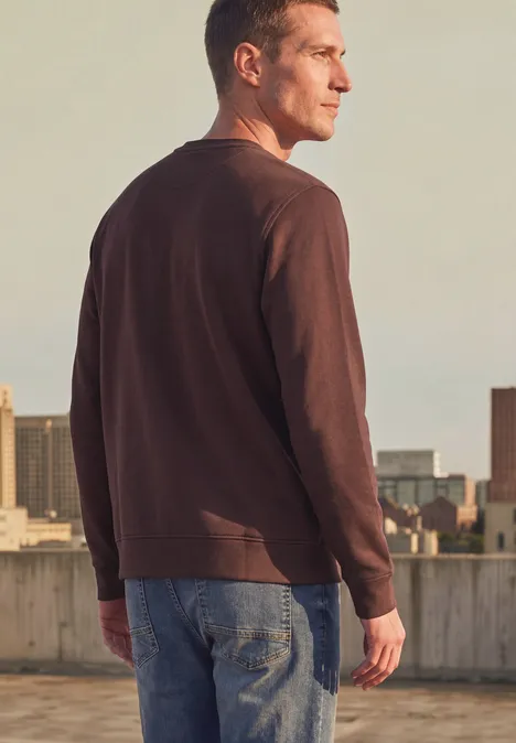 Homme en t-shirt à manches longues marron et jean se tient sur un toit, regard vers la ville.