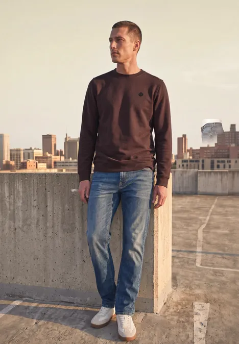 Un homme sur un toit-terrasse regarde la ville, vêtu d’un sweat marron, d’un jean et de baskets blanches.