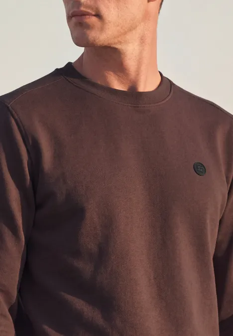 Un homme porte un sweat-shirt marron uni avec un petit logo rond sur la poitrine.