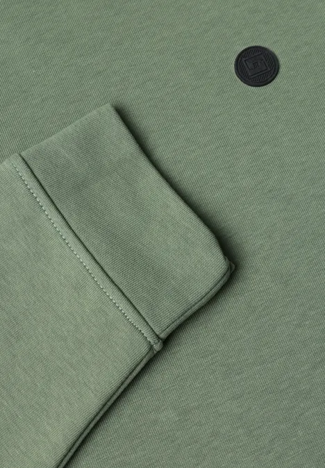 Nahansicht eines grünen Kleidungsstücks mit aufgesetzter Tasche und rundem schwarzen Logo.