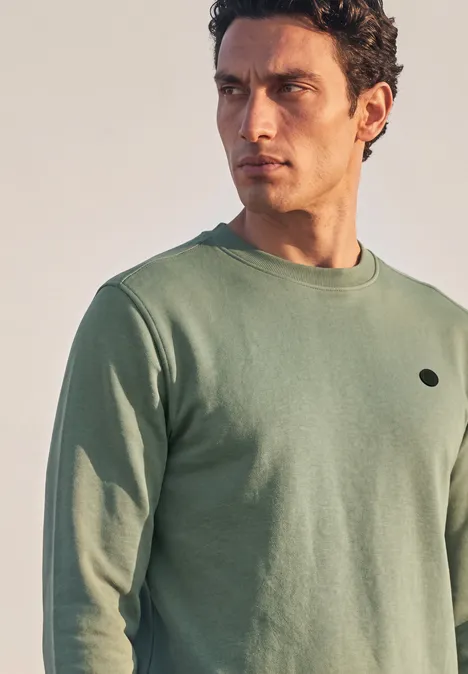 Ein Mann mit lockigem Haar trägt ein schlichtes, hellgrünes Sweatshirt mit Rundhalsausschnitt.