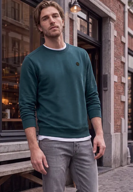 Un homme en sweat vert et jean gris pose détendu devant une boutique sur une rue pavée.