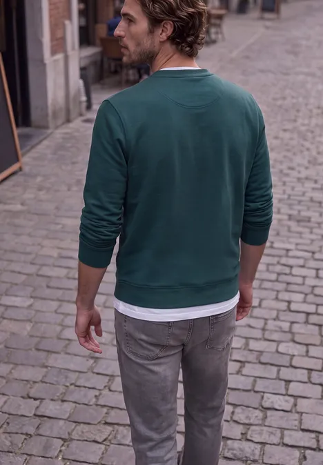 Un homme de dos en pull vert et pantalon gris sur une rue pavée.