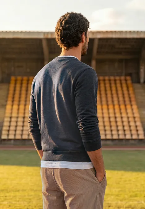 Un homme en pull se tient sur la pelouse d’un stade, dos tourné vers les tribunes vides.