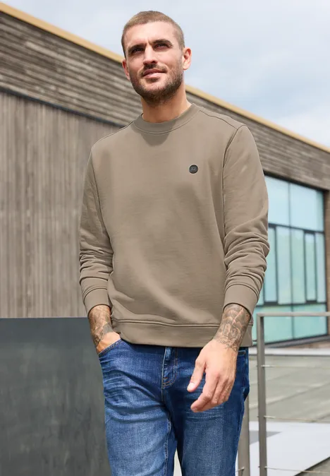 Un homme décontracté en pull olive et jean bleu pose devant un bâtiment moderne.