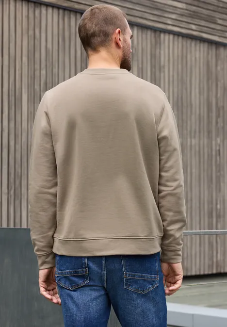 Un homme de dos portant un sweat gris-brun devant un mur en bois.