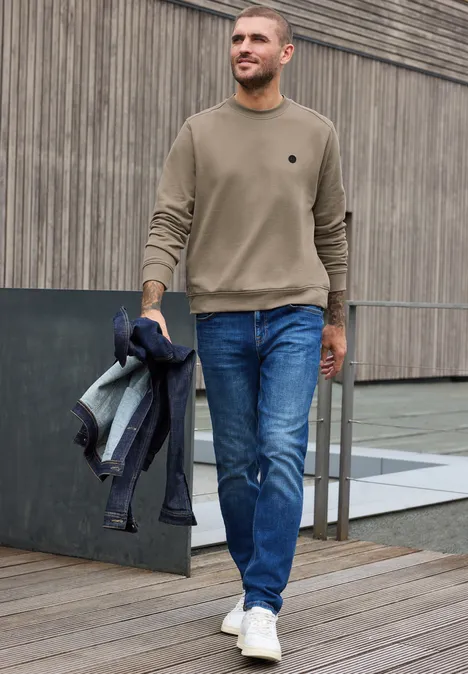 Un homme marche en jeans, pull beige et baskets blanches, une veste à la main.