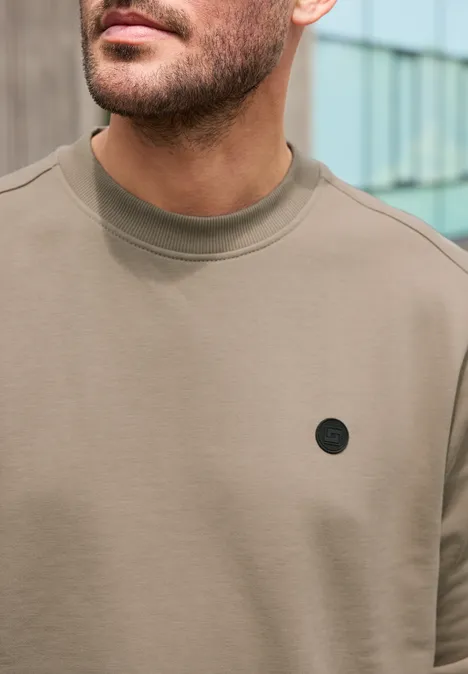 Gros plan d’un homme en t-shirt taupe à col rond, orné d’un petit logo noir sur la poitrine.