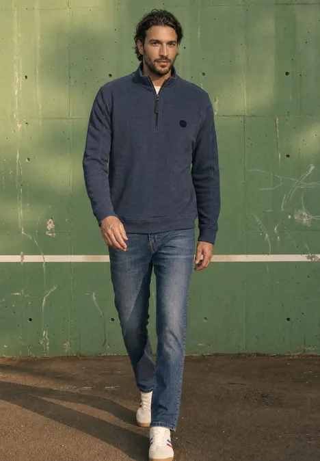 Ein Mann in Jeans, weißen Turnschuhen und blauem Zip-Pullover geht vor einer grünen Wand mit weißer Linie.