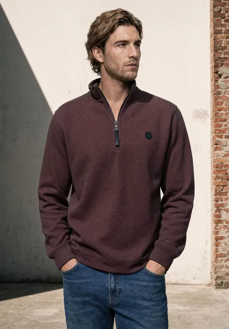 Ein Mann trägt einen bordeauxroten Pullover mit halbem Reißverschluss und steht vor einer Backsteinwand.