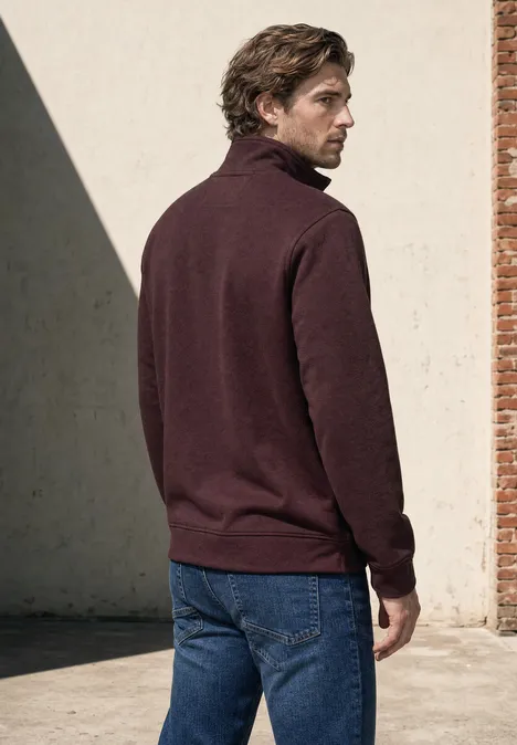 Mann mit braunem Rollkragenpullover und Jeans steht seitlich vor einer Wand mit einem Ziegelpfeiler.