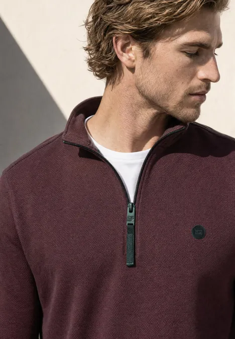 Ein Mann in einem burgunderroten Pullover mit Reißverschluss und kleinem Logo, der nach unten blickt.