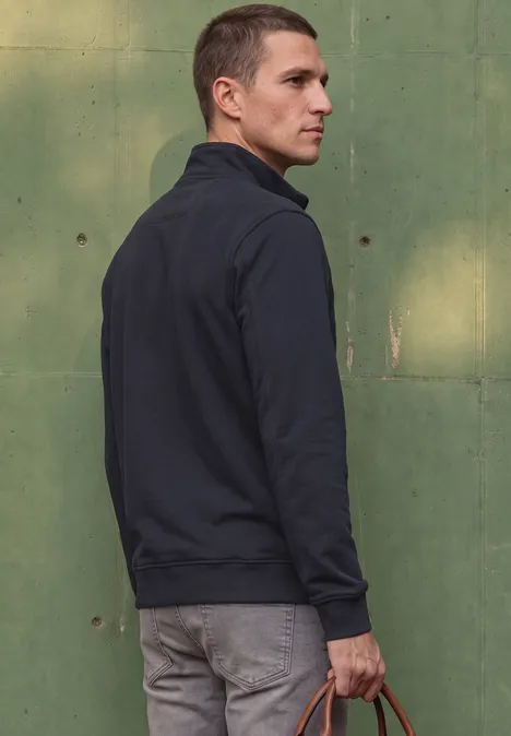 Ein Mann in dunkler Jacke und grauer Hose steht seitlich vor einer grünen Wand und hält eine Tasche.