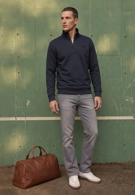 Ein Mann in marineblauem Pullover, grauer Hose und weißen Sneakers steht neben brauner Reisetasche vor grünem Holz.