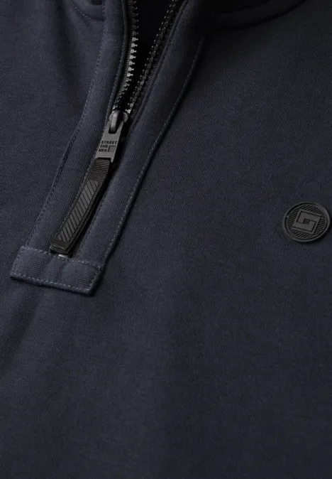 Nahaufnahme eines dunkelblauen Sweatshirts mit kurzem Reißverschluss und kleinem runden Logo auf der Brust.