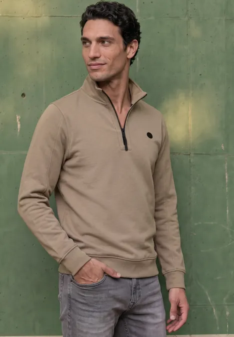 Ein Mann steht vor einer grünen Wand, trägt einen beigen Pullover mit kurzem Reißverschluss und Jeans.