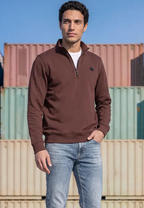 Ein Mann in braunem Troyer und Jeans steht vor bunten Containerwänden bei klarem Himmel.