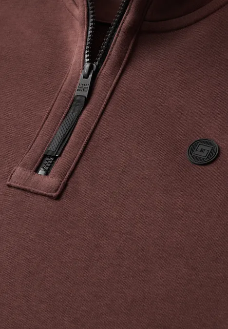 Braunes Sweatshirt mit halbem Reißverschluss und kleinem, rundem Logo auf der Brust.