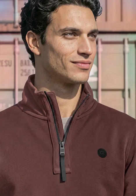 Ein Mann in einem dunkelroten Zip-Pullover vor bunten Containern.