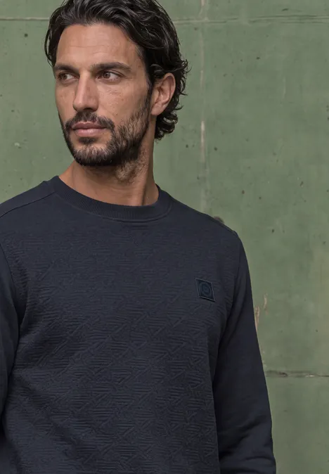 Ein Mann steht in dunklem Pullover vor einer grünen Wand und schaut nach rechts.
