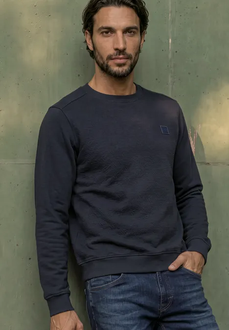 Ein Mann steht vor einer grünen Wand, trägt einen dunkelblauen Pullover und blaue Jeans.