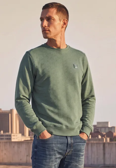 Ein Mann steht draußen an einem Geländer, trägt grünes Sweatshirt und Jeans.