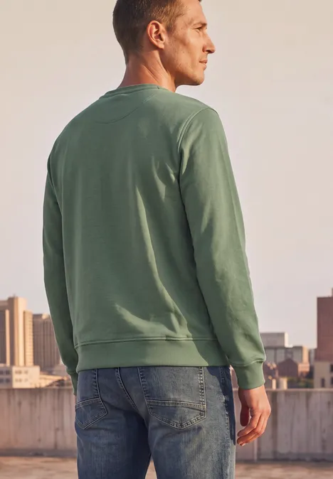 Mann von hinten trägt grünes Sweatshirt und Jeans, steht auf Dach mit Stadtkulisse.