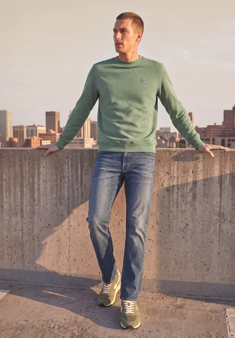 Ein Mann in grüner Sweatshirt und Jeans steht auf einem Dach und schaut über die Stadt.