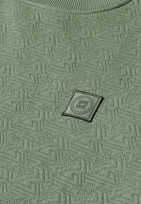 Grüner Stoff mit geometrischem Allover-Muster und kleinem, quadratischen Logo-Patch.