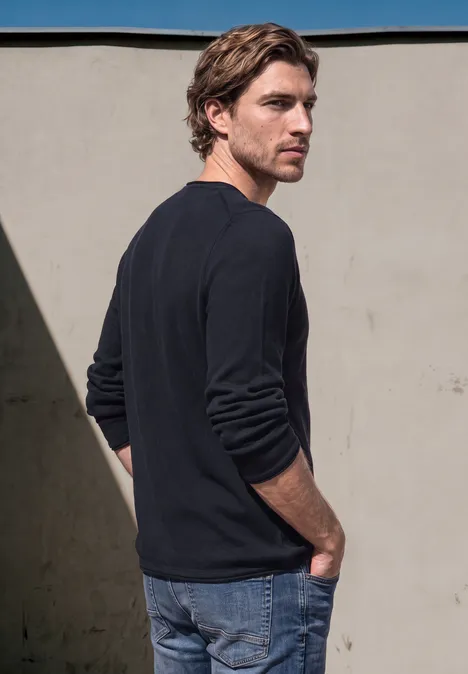 Ein lässiger Mann in dunklem Pullover steht seitlich, Hände in den Hosentaschen vor einer hellen Wand.