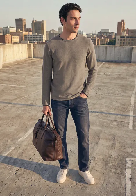 Mann in Jeans und grauem Pullover steht auf Parkhausdach, trägt braune Tasche, Stadtsilhouette im Hintergrund.