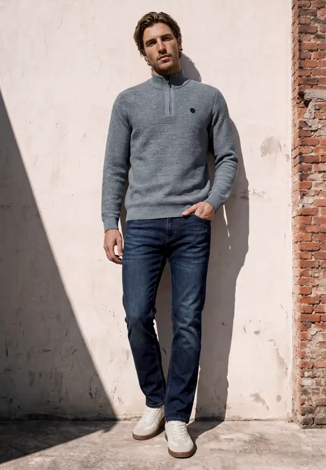 Pullover mit Troyerkragen und Struktur vintage blue Ein Mann lehnt entspannt an einer sonnenbeschienenen Wand, trägt grauen Pullover, blaue Jeans und helle Sneaker.