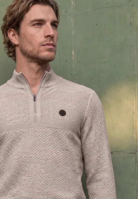 Ein Mann in einem hellbeigen Pullover mit Reißverschluss und Strukturmuster vor grüner Metallwand.