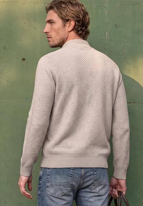 Ein Mann in grauem Strickpullover und Jeans steht vor einer grünen Wand und hält eine Jacke in der Hand.