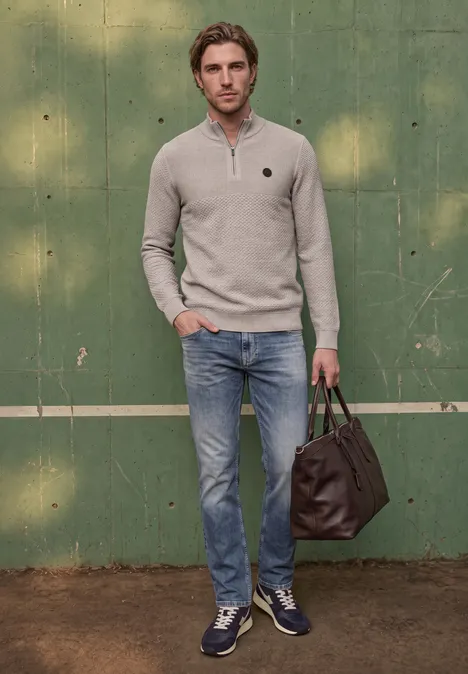 Ein Mann trägt einen hellgrauen Pulli, Jeans, blaue Sneaker und hält eine braune Tasche vor grüner Wand.