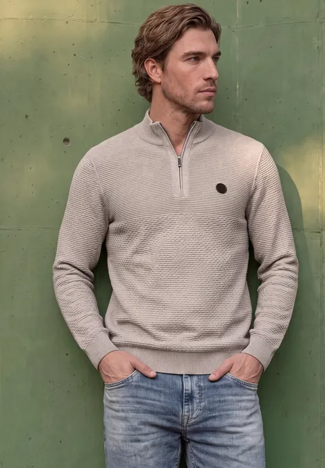 Ein Mann trägt einen beigen Pullover mit Reißverschluss und Jeans vor grünem Hintergrund.