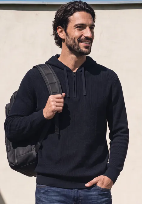 Ein lächelnder Mann trägt einen dunklen Hoodie und Rucksack vor neutraler Wand.