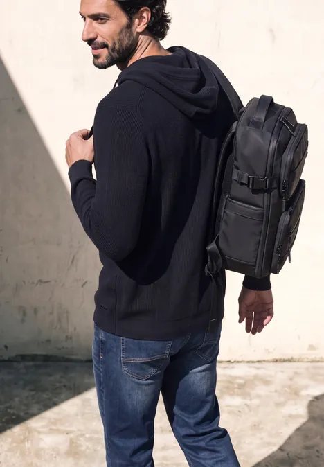 Ein Mann in schwarzer Kapuzenjacke und Jeans trägt einen schwarzen Rucksack und geht an einer hellen Wand vorbei.