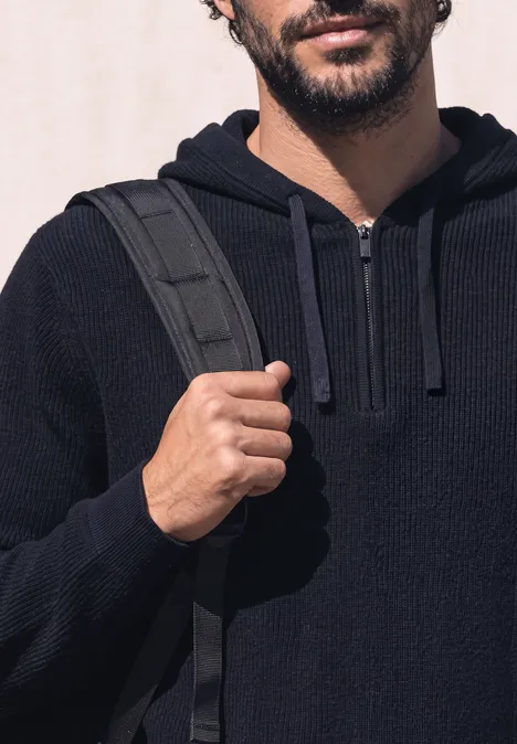 Ein Mann trägt einen dunkelblauen Kapuzenpullover und hält den Rucksackgurt auf der Schulter