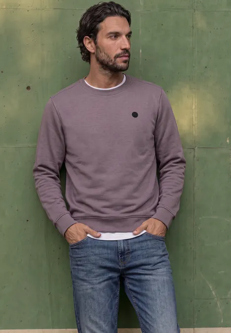 Ein Mann in lila Pullover und Jeans steht vor einer grünen Wand mit Händen in den Taschen.