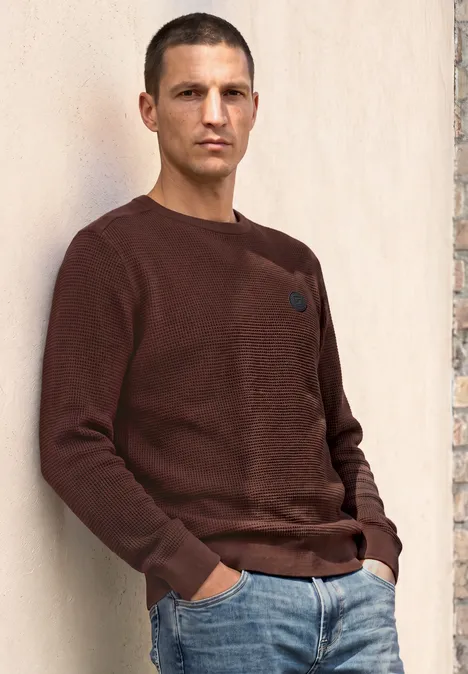 Ein Mann lehnt an einer Wand, trägt einen braunen Pullover und Jeans.