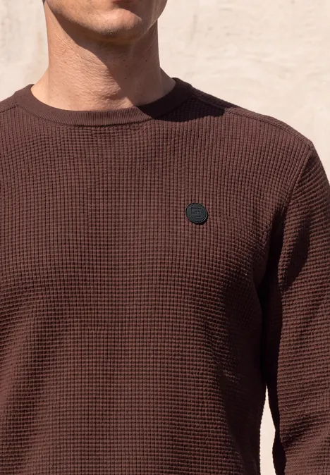 Ein Mann trägt ein braunes, strukturiertes Shirt mit kleinem schwarzem Badge.